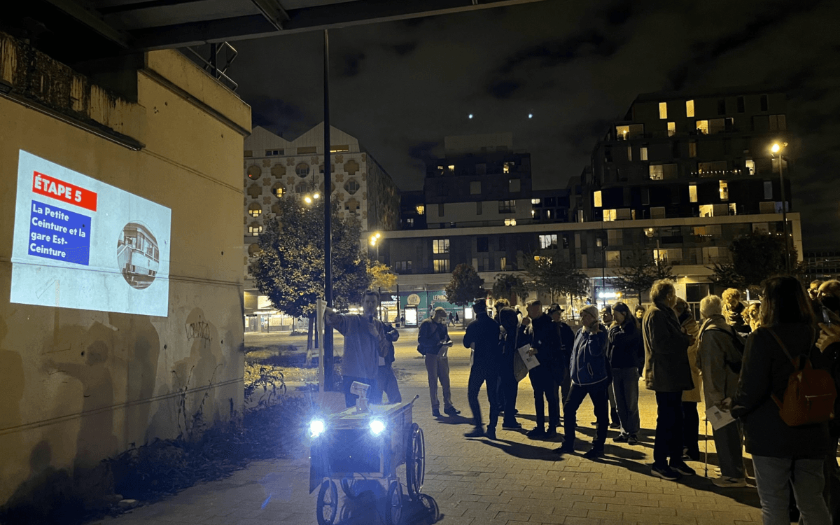 La promenade réunit un groupe d’une trentaine de personnes, guidé à travers le quartier par une charrette mobile équipée d’un picoprojecteur. À chaque étape, les façades deviennent des supports de projection pour faire apparaître photographies d’archives, images animées et comparaisons entre passé et présent.