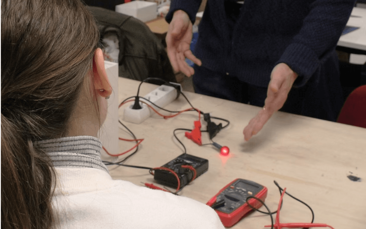 Photographie d'une participante à l'atelier brico électro vue de dos. Matériels électroniques et mains de l'animateur visibles sur la table à laquelle elle est assise.